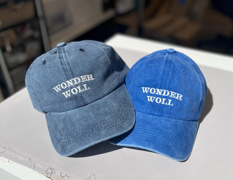 May include: Two blue baseball caps. One is a faded denim blue, the other a bright blue. Both caps feature the white embroidered text "WONDER WOLL" across the front. A casual accessory for everyday wear.