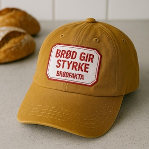 Può includere: Un berretto da baseball giallo senape con una toppa ricamata in rosso e bianco che recita "Brod Gir Styrke Brodfakta".