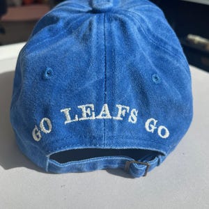 May include: A blue baseball cap with the words "GO LEAFS GO" embroidered in white. The cap has a curved brim and an adjustable strap at the back. The cap is made of a soft, textured fabric.