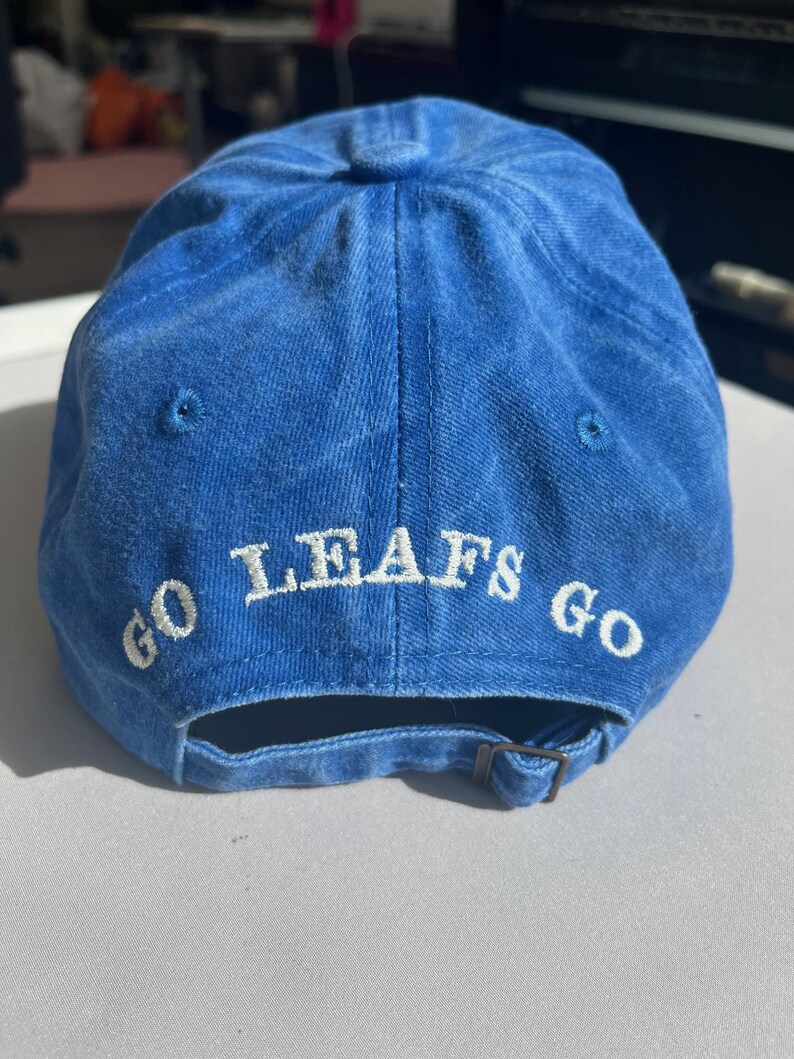 May include: A blue baseball cap with the words "GO LEAFS GO" embroidered in white. The cap has a curved brim and an adjustable strap at the back. The cap is made of a soft, textured fabric.