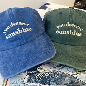 May include: Two baseball caps, one blue and one green, are displayed. Both caps have the phrase "you deserve sunshine" embroidered in white. The caps are made of a soft, washed fabric.