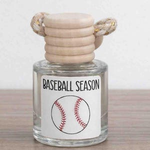 May include: A clear glass bottle with a wooden cap and a label that reads "BASEBALL SEASON." The label features a baseball graphic with red stitching. The cap has a decorative woven detail.