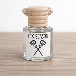May include: A clear glass bottle with a wooden cap and a white label. The label reads "LAX SEASON" above a black graphic of crossed lacrosse sticks. The bottle has a beige rope detail at the top.