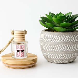May include: A clear glass car air freshener with a wooden top and a rope. The label reads "BEACH BABE" with sunglasses. It sits on a wooden base next to a potted succulent.