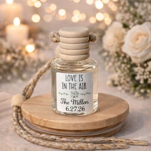 May include: A clear glass bottle with a wooden cap and a label that reads "LOVE IS IN THE AIR" with the names "The Millers" and the date "6.27.26". The bottle sits on a wooden base with a rope detail, with a blurred background of lights and flowers.