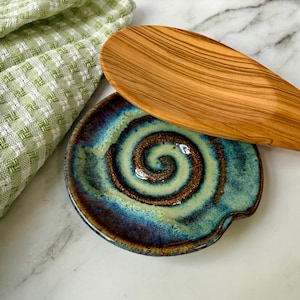 May include: A ceramic spoon rest with a blue, brown, and green spiral design. A wooden spoon rests on the spoon rest. A green and white checkered kitchen towel is in the background.