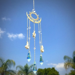 May include: A celestial suncatcher featuring a gold sun and crescent moon design. Clear crystal beads and teardrop prisms hang from fine chains. The suncatcher is suspended against a blue sky with clouds and greenery in the background.