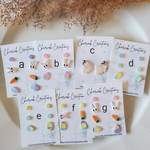 May include: Six sets of Easter-themed polymer clay earrings. Each set includes three pairs of earrings with different designs, including bunnies, carrots, and eggs. The earrings are displayed on white cards with the text "Charcob Creations" and their website address.