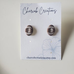May include: A pair of brown and white football-shaped earrings on a white card with the text "Charcob Creations" and "charcobcreations.etsy.com".