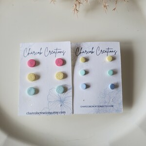 May include: Two sets of colourful stud earrings on white cards. The first set has pink, yellow, and light blue earrings. The second set has yellow, light blue, and dark blue earrings. The cards have the text "Charcob Creations" and a website address.