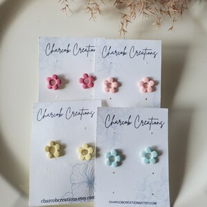 May include: Four pairs of flower-shaped stud earrings on white cards. The earrings are in pink, light pink, yellow, and light blue.