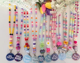 May include: A collection of eleven beaded lanyards featuring colorful silicone beads in pastel and bright hues. Each lanyard includes a unique charm, such as flowers, books, or hearts, and a circular badge personalized with the name Ms. or Miss Reyes.