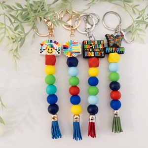 May include: Four colorful beaded keychains with different autism awareness charms. The charms include a smiley face with the words "autism mom", a sunflower, a black charm with the text "My child may be non-verbal but this mama ain't", and a butterfly with the words "Autism Awareness".