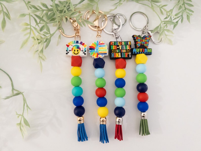 May include: Four colorful beaded keychains with different autism awareness charms. The charms include a smiley face with the words "autism mom", a sunflower, a black charm with the text "My child may be non-verbal but this mama ain't", and a butterfly with the words "Autism Awareness".