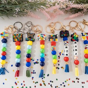 May include: Seven colorful beaded keychains with different autism awareness charms. The charms include a puzzle piece, a heart with the words "I Love Autism", a smiley face, a butterfly, and a charm with the text "My Child May Be Non-Verbal But This Home Ain't Quiet". The keychains have blue, red, yellow, and green beads.