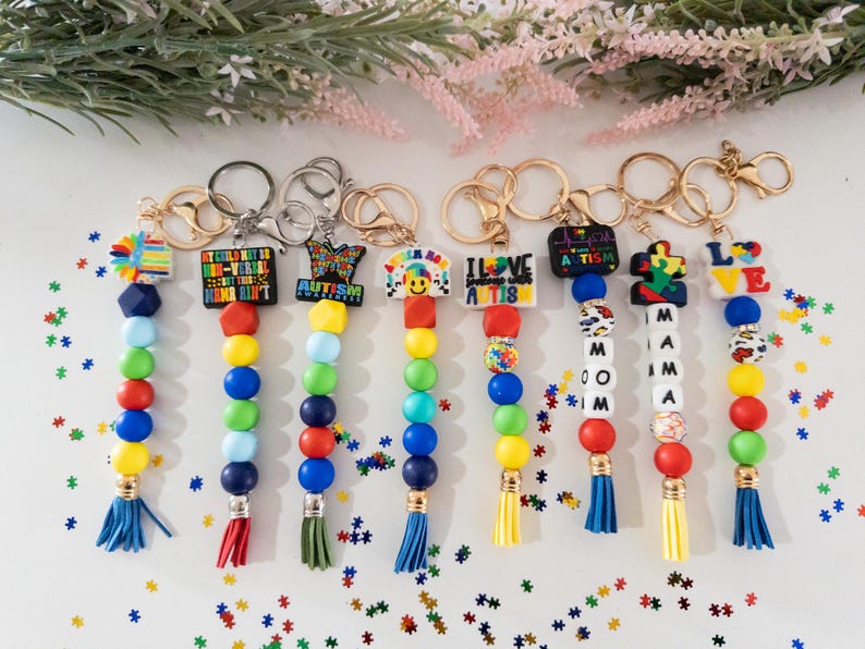 May include: Seven colorful beaded keychains with different autism awareness charms. The charms include a puzzle piece, a heart with the words "I Love Autism", a smiley face, a butterfly, and a charm with the text "My Child May Be Non-Verbal But This Home Ain't Quiet". The keychains have blue, red, yellow, and green beads.