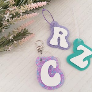 May include: Three letter-shaped keychains and tags. The letter "C" is attached to a silver keychain. The letters "R" and "Z" are on tags. The letters are white, with a glittery, colorful border. A sprig of flowers is in the background.