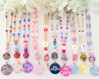 May include: A collection of colorful beaded lanyards with various charms, including apples, flowers, and musical notes. Each lanyard features a teacher's name on a round pendant. The lanyards are adorned with wooden and colorful beads.