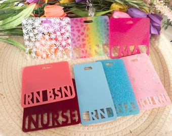 May include: A collection of rectangular acrylic badge reels featuring various designs including white snowflakes, rainbow leopard print, solid pink glitter, solid coral, light blue, blue glitter, and light pink with cut-out text like RN, BSN, and NURSE.