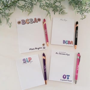 May include: White notepads with colorful text, including "BCBA," "SLP," and "OT Occupational Therapist." Each notepad has a matching pen. The image is styled with greenery.