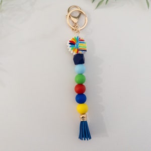 May include: A colorful beaded keychain with a blue tassel. The keychain features a small, white charm with a colorful flower design. The beads are blue, green, red, and yellow.