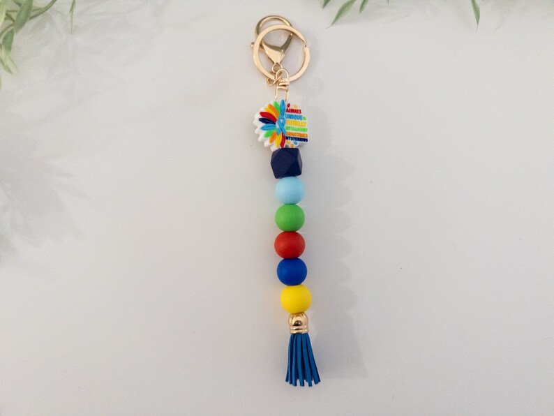 May include: A colorful beaded keychain with a blue tassel. The keychain features a small, white charm with a colorful flower design. The beads are blue, green, red, and yellow.