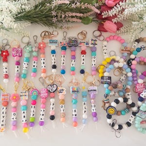 May include: A collection of beaded keychains and wristlets featuring colorful silicone and plastic beads, metal key rings, and various charms with text including SLP, OT, ABA, and other phrases. The items are arranged in a group on a flat surface.