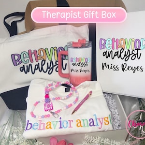 May include: A gift box containing a white tote bag, a white tumbler with a pink handle, a white notebook, a white sweatshirt, a beaded lanyard, and a pink pen. The items feature the text Behavior Analyst and Miss Reyes in colorful fonts.