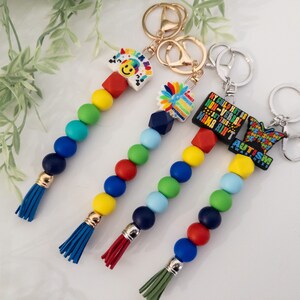 May include: Four colorful beaded keychains with different charms. The charms include a rainbow with a smiley face, a flower, a black charm with the text "My child may be non-verbal but this mama ain't autism awareness", and a blue butterfly with the text "Autism Awareness".