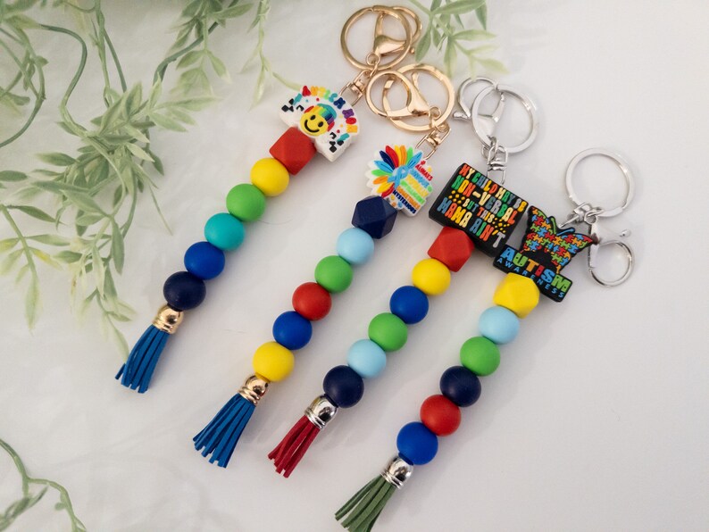 May include: Four colorful beaded keychains with different charms. The charms include a rainbow with a smiley face, a flower, a black charm with the text "My child may be non-verbal but this mama ain't autism awareness", and a blue butterfly with the text "Autism Awareness".
