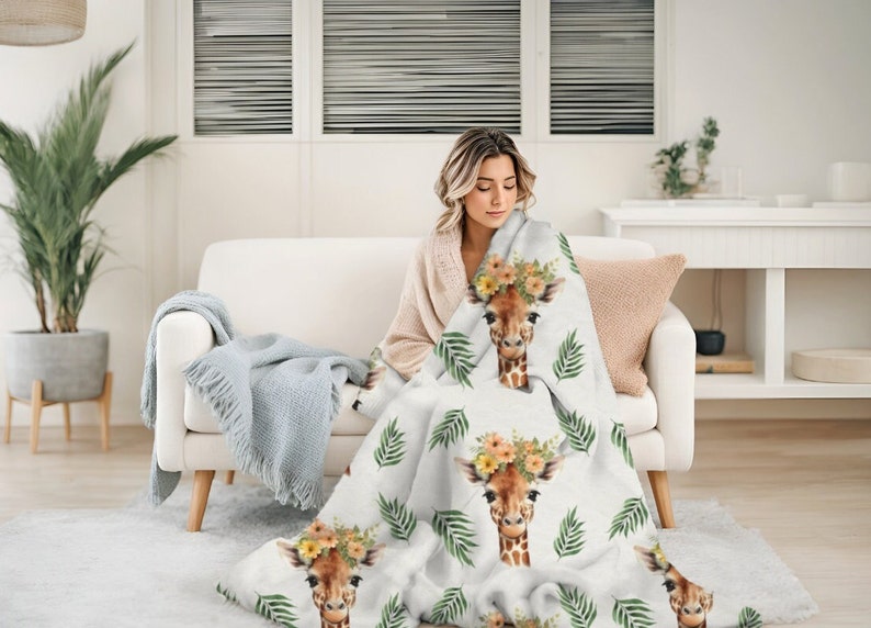 May include: A white throw blanket with a repeating pattern of giraffes wearing flower crowns and green palm leaves. The blanket is draped over a white couch with a person sitting on it.
