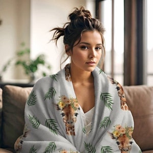 May include: A soft, gray fleece blanket with a repeating pattern of giraffes wearing flower crowns and green palm leaves. The blanket is draped over a person sitting on a couch.