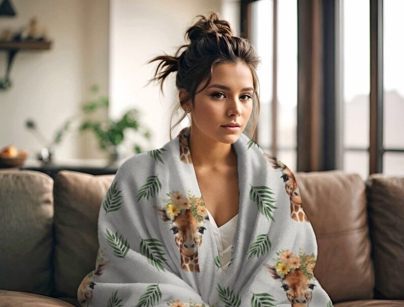 May include: A soft, gray fleece blanket with a repeating pattern of giraffes wearing flower crowns and green palm leaves. The blanket is draped over a person sitting on a couch.