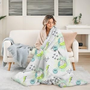 Pode incluir: Uma manta de lã branca com um padrão repetido de bolas de voleibol verde e azul, rostos sorridentes, corações e raios. A manta está drapeada sobre um sofá branco.