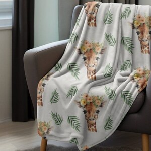 May include: A white throw blanket with a repeating pattern of giraffes wearing flower crowns and green palm leaves.