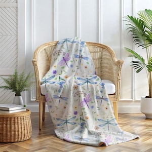 Pode incluir: Uma manta branca com um padrão repetido de libélulas azuis e roxas e flores de aquarela. A manta está drapeada sobre uma cadeira de vime.