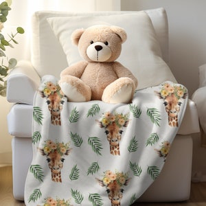 May include: White plush throw blanket with a repeating pattern of giraffe faces wearing floral crowns and green palm leaves. A brown teddy bear is sitting on the blanket.