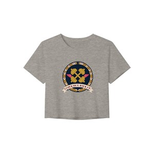 Könnte beinhalten: Ein graues, kurzes T-Shirt mit einem kreisförmigen Aufdruck auf der Brust. Das Design zeigt eine goldene Hantel mit kleinen rosa Flügeln, umgeben von einem goldenen Weizenkranz auf dunkelblauem Grund, mit einem Banner mit der Aufschrift CLOCK OUT. RACK UP.