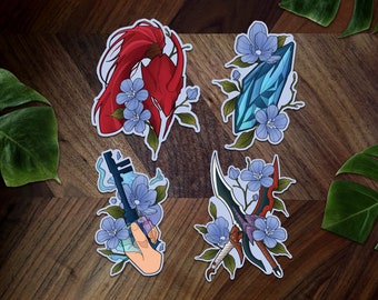 May include: Four fantasy-themed sticker designs. The stickers show a red helmet, a blue crystal, a key held by a hand, and a sword, each decorated with blue flowers and green leaves. The stickers are on a wooden surface.