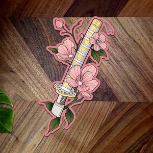 May include: A pink and yellow sticker with a stylized illustration of a katana sword wrapped in a floral design. The sword is surrounded by pink flowers and green leaves.