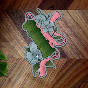 May include: A green and white floral design with a pink ribbon. The design features a green cylinder with a white flower and a pink ribbon wrapped around it.