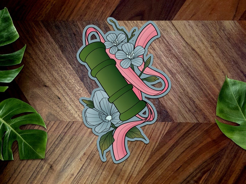 May include: A green and white floral design with a pink ribbon. The design features a green cylinder with a white flower and a pink ribbon wrapped around it.