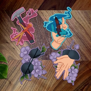 May include: Four colorful stickers with different designs. The first sticker is pink and features a hammer, a voodoo doll, and needles. The second sticker is blue and features a hand holding a blue flame. The third sticker is purple and features a pair of sunglasses with flowers. The fourth sticker is purple and features two hands holding each other with flowers.