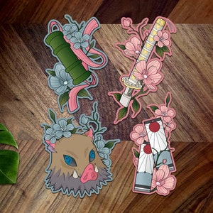 Może przedstawiać: Cztery kolorowe naklejki przedstawiające postacie i symbole z anime Demon Slayer. Naklejki przedstawiają zieloną bambusową rurkę, miecz z żółtą rękojeścią, maskę dzika i kratkowany wzór z czerwonym słońcem.