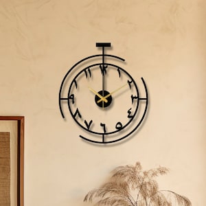 May include: A black metal clock with Roman numerals and gold hands hanging on a beige wall. The clock is in a living room setting with a wooden cabinet, a vase with dried flowers, and a small brown pot.