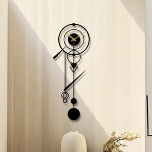 Op de afbeelding: Een geometrische wandklok in zwart en goud met een cirkelvormig ontwerp en een lange, dunne slinger. De klok hangt aan een lichtbruine muur in een modern huis.