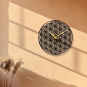 Può includere: Un orologio da parete nero e dorato con un design a fiore della vita. L'orologio è appeso a una parete color marrone chiaro con una panca di legno in primo piano.