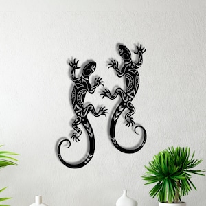 Op de afbeelding: Zwarte metalen wandkunst met twee gestileerde hagedissen in een symmetrisch ontwerp. De hagedissen hebben ingewikkelde patronen en zijn op een witte muur gemonteerd. Het kunstwerk voegt een decoratief tintje toe aan elke interieurruimte.