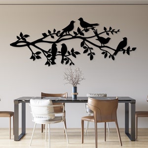 May include: Black metal wall art depicting birds perched on a tree branch with leaves. The artwork is mounted on a neutral-colored wall above a dining table with chairs. The birds are silhouetted against the wall.
