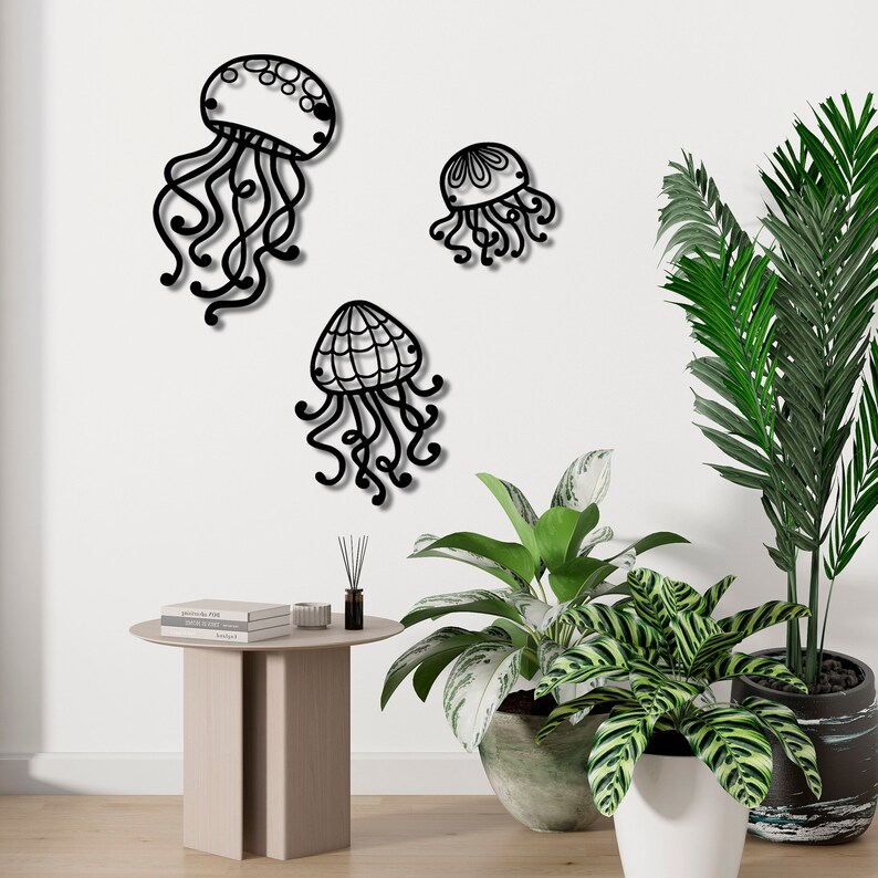 May include: Three black metal wall decor pieces shaped like jellyfish with intricate designs. The jellyfish are hanging on a white wall with two potted plants in the foreground.